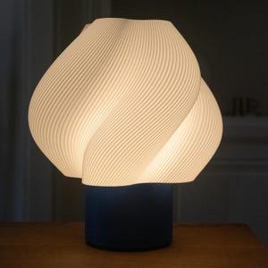 May include: A table lamp featuring a white, ribbed, spiraling shade with a distinct organic, bulbous shape. The shade is illuminated from within, casting a soft glow. It is supported by a solid, dark blue cylindrical base that provides a contrast.