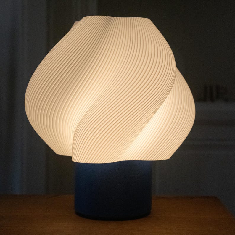 May include: A table lamp featuring a white, ribbed, spiraling shade with a distinct organic, bulbous shape. The shade is illuminated from within, casting a soft glow. It is supported by a solid, dark blue cylindrical base that provides a contrast.