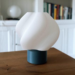 May include: A table lamp featuring a white, spiraling, ribbed shade with a textured surface. The shade is mounted on a solid, cylindrical base in a deep teal blue color. The lamp has a rounded, bulbous silhouette and a smooth, matte finish.