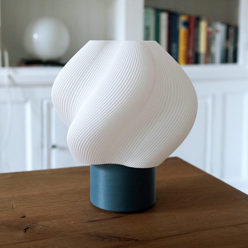 May include: A table lamp featuring a white, spiraling, ribbed shade with a textured surface. The shade is mounted on a solid, cylindrical base in a deep teal blue color. The lamp has a rounded, bulbous silhouette and a smooth, matte finish.
