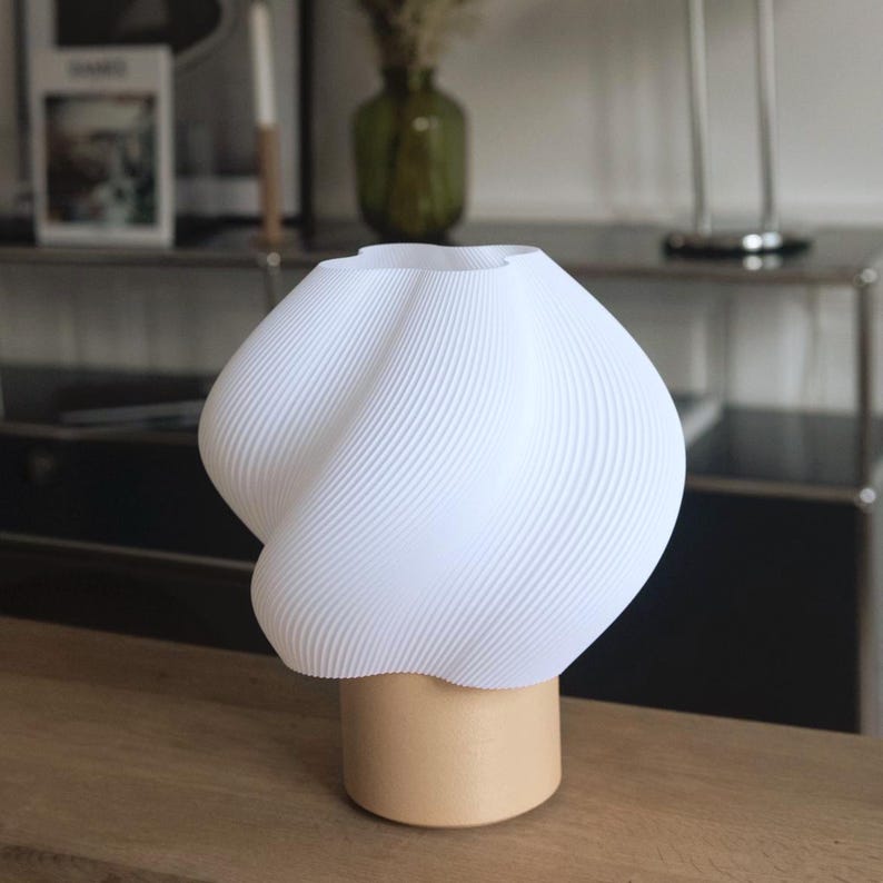 May include: A table lamp featuring a white, 3D-printed shade with a distinct twisted, ribbed texture. The shade is mounted on a cylindrical, light-colored wooden base. The overall design is rounded and bulbous, providing a clean, neutral aesthetic.