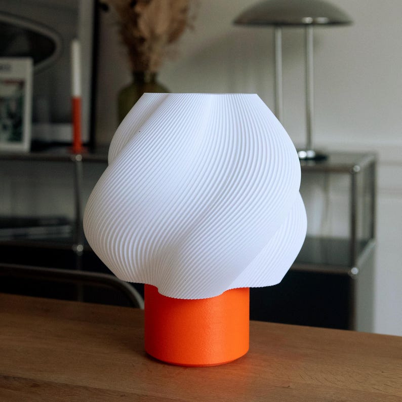 May include: A table lamp featuring a white, 3D-printed shade with a distinct twisted, ribbed texture. The shade is supported by a solid, cylindrical orange base. The lamp has a smooth, matte finish and a compact, balanced structural design.