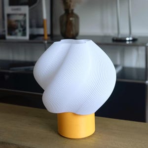 May include: A table lamp featuring a white, spiraled, ribbed shade with a distinct twisted, organic shape. The base of the lamp is a solid, cylindrical, matte yellow component that provides a stable foundation for the textured white top.