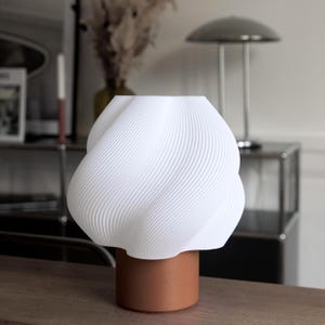 May include: A table lamp featuring a white, spiraled, textured shade with a wavy, organic form. The shade is supported by a solid, cylindrical, terracotta-colored base. The lamp has a smooth, matte finish and a distinct, layered, 3D-printed appearance.