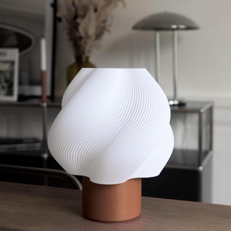 May include: A table lamp featuring a white, spiraled, textured shade with a wavy, organic form. The shade is supported by a solid, cylindrical, terracotta-colored base. The lamp has a smooth, matte finish and a distinct, layered, 3D-printed appearance.