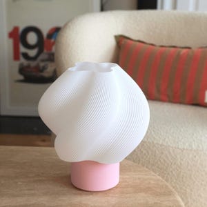 May include: A table lamp featuring a white, ribbed, twisted shade with a rounded, bulbous form. The shade is supported by a solid, cylindrical base in a matte light pink finish. The lamp has a distinct, textured surface on the shade.