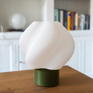 May include: A table lamp featuring a white, spiraled, ribbed shade with a rounded, organic shape. The shade is supported by a solid, cylindrical base in a matte olive green finish. The lamp has a clean, structured design with visible texture.