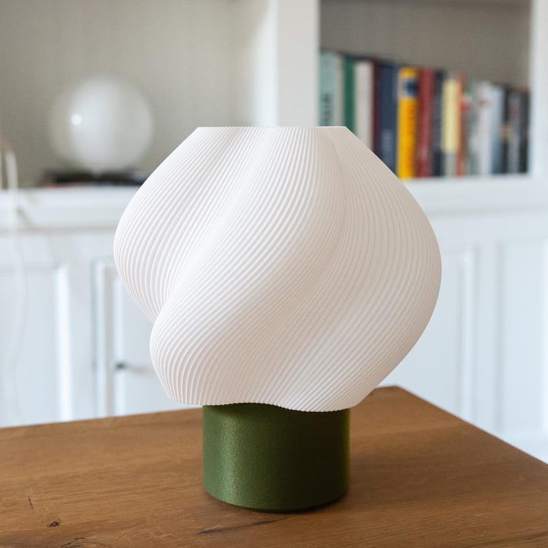 May include: A table lamp featuring a white, spiraled, ribbed shade with a rounded, organic shape. The shade is supported by a solid, cylindrical base in a matte olive green finish. The lamp has a clean, structured design with visible texture.