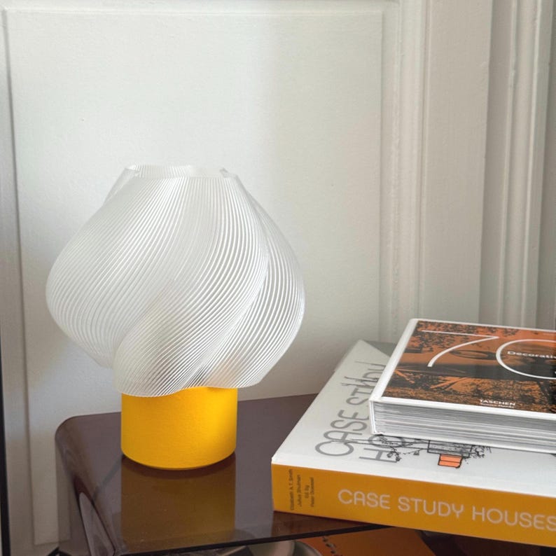 May include: A table lamp featuring a translucent white, spiraled, ribbed shade with a rounded, bulbous form. The base of the lamp is a solid, cylindrical, bright yellow block. The lamp sits next to a book titled Case Study Houses.