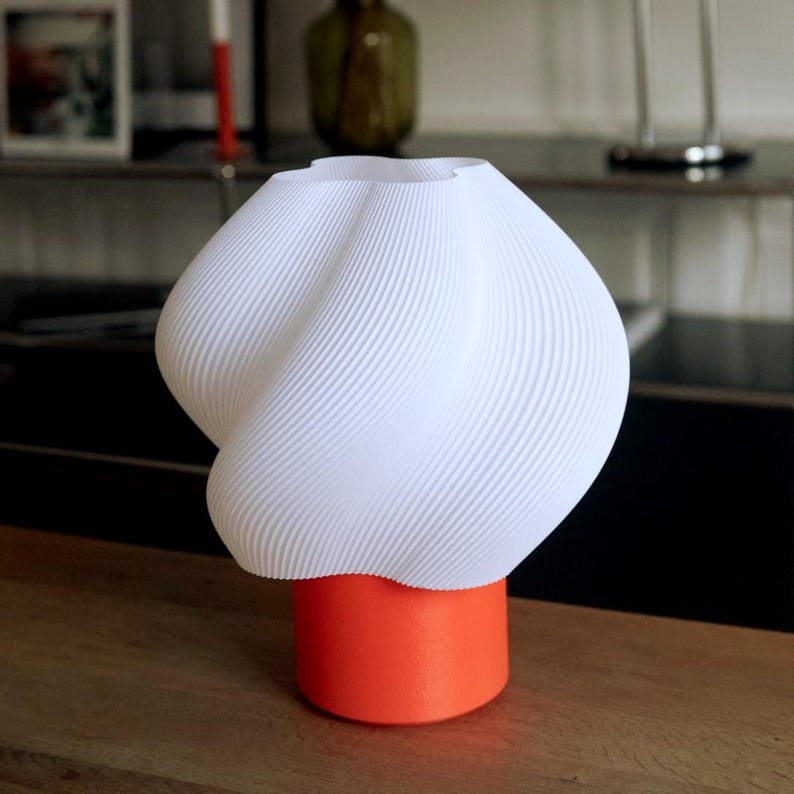 May include: A table lamp featuring a white, ribbed, twisted shade with a wavy top edge. The shade is supported by a solid, cylindrical, bright orange base. The design incorporates a textured surface on both the shade and the base, creating a distinct contrast between the white upper section and the vibrant orange lower section.