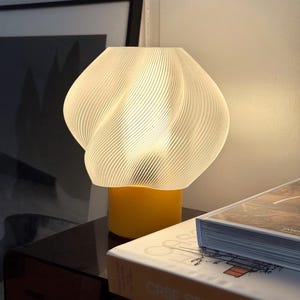 May include: A table lamp featuring a translucent white, spiraled, ribbed shade with a soft glow. The lamp has a solid, cylindrical yellow base that supports the textured, organic-shaped shade, creating a warm and diffused light effect.