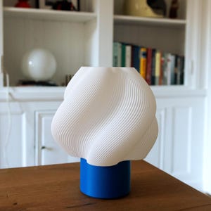May include: A table lamp featuring a white, spiraled, textured shade with a wavy, ribbed surface. The shade is supported by a solid, cylindrical, matte blue base. The lamp has a distinct, modern, geometric silhouette with a wide top and narrow base.