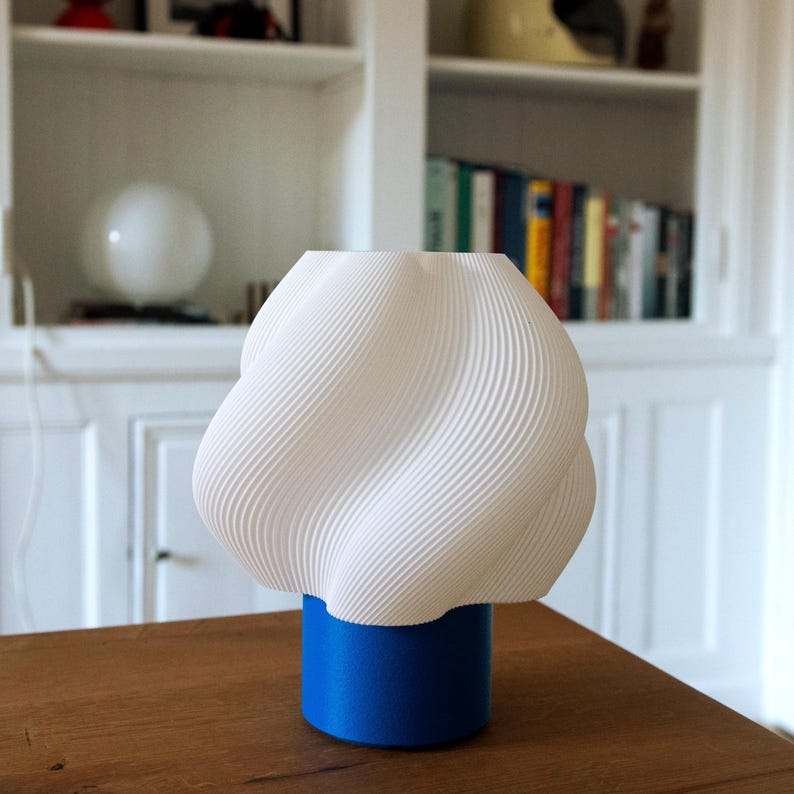 May include: A table lamp featuring a white, spiraled, textured shade with a wavy, ribbed surface. The shade is supported by a solid, cylindrical, matte blue base. The lamp has a distinct, modern, geometric silhouette with a wide top and narrow base.