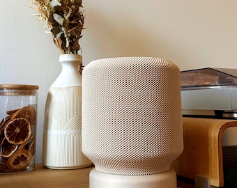 以下が含まれることがあります： オフホワイトのテクスチャ加工されたファブリックメッシュの外装と頑丈なベースを備えた円筒形のスマートスピーカー。スピーカーは丸みを帯びた上端と、表面全体に均一なドットパターンが施されており、独立した電子機器として直立しています。