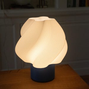 May include: A table lamp featuring a white, spiraled, textured shade with a wavy, organic form that emits a warm glow. The shade is supported by a solid, cylindrical, dark blue base that provides a stable foundation for the light fixture.
