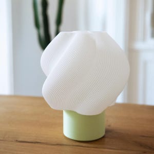 May include: A table lamp featuring a white, ribbed, twisted shade with a rounded, organic shape. The shade is supported by a solid, cylindrical base in a soft, light green color. The lamp has a smooth, matte finish throughout its structure.