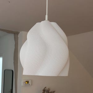 Puede incluir: Lámpara colgante blanca y moderna con un diseño ondulado único. La lámpara cuelga de un cable blanco y presenta una superficie texturizada con líneas verticales. Cuelga en una habitación con paredes de colores neutros.