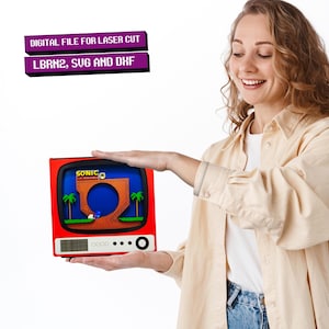 May include: A red and black retro television with a screen showing a pixelated image of Sonic the Hedgehog running through a level. The text "SONIC THE HEDGEHOG" is visible on the screen. The television is being held by a person's hands.