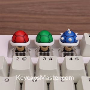 May include: Three keycaps featuring different colored Super Mario Bros. turtle shells. The red shell is on the left, the green shell is in the middle, and the blue spiked shell is on the right. The keycaps are on a white keyboard with black keys.