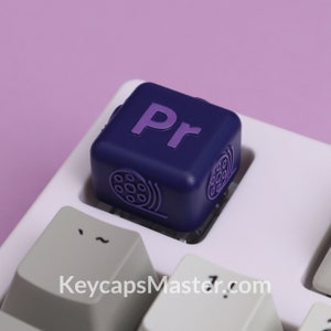 May include: A purple and blue keycap with the letters "Pr" and a film reel symbol. The keycap is on a white keyboard with the text "KeycapsMaster.com" visible.