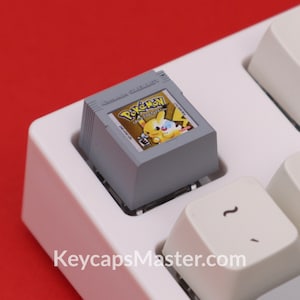 May include: A gray and white keyboard keycap with a miniature Game Boy cartridge featuring the Pokemon Yellow game. The cartridge is labeled "Nintendo Game Boy" and "Pokemon Yellow Special Pikachu Edition".