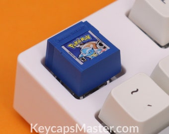 May include: A blue and white Pokemon Game Boy cartridge keycap for a mechanical keyboard. The keycap features the text "Pokemon Game Boy" and an image of a Pokemon character.