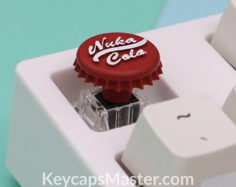 Pode incluir: Um keycap de teclado mecânico vermelho e branco em forma de tampa de garrafa Nuka Cola. O keycap tem a forma de uma tampa de garrafa e tem o logotipo da Nuka Cola.