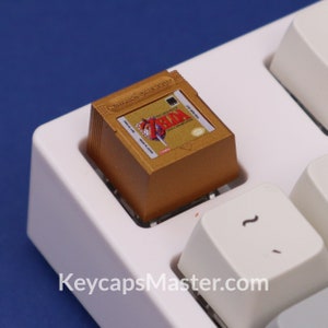 May include: A gold-colored Nintendo Game Boy cartridge with a label featuring the image of Link from The Legend of Zelda: A Link to the Past. The cartridge is sitting on a white keyboard.