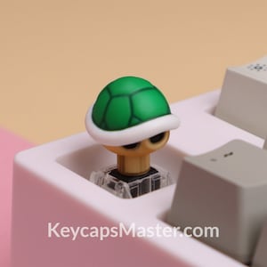 Peut inclure: Un capuchon de touche de clavier mécanique en forme de carapace de tortue verte et blanche. Le capuchon de touche est en plastique et présente un design réaliste. Le capuchon de touche est posé sur un clavier blanc.