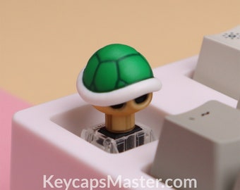 May include: A green and white turtle shell keycap for a mechanical keyboard. The keycap is made of plastic and has a realistic design. The keycap is sitting on a white keyboard.