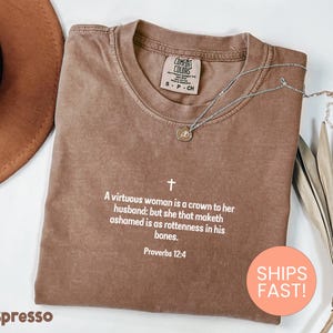 May include: A brown short-sleeved t-shirt with a crew neckline featuring white printed text that reads: A virtuous woman is a crown to her husband: but she that maketh ashamed is as rottenness in his bones. Proverbs 12:4, with a small cross symbol.