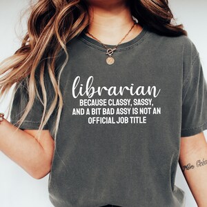 May include: A charcoal grey short-sleeve t-shirt featuring white text that reads: librarian BECAUSE CLASSY, SASSY, AND A BIT BAD ASSY IS NOT AN OFFICIAL JOB TITLE. The shirt has a crew neckline and a relaxed fit with a vintage-style washed finish.