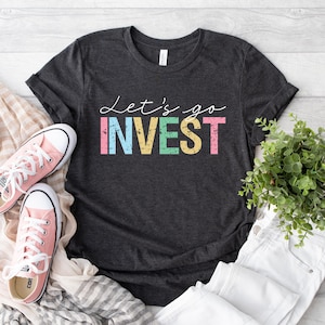 Könnte beinhalten: Ein dunkel anthrazitfarbenes Kurzarm-T-Shirt mit dem weißen Schreibschrift-Text Let's go und dem in Blockbuchstaben geschriebenen Wort INVEST in abgenutzten Pastellfarben wie Rosa, Blau, Gelb und Grün. Das T-Shirt wird mit rosa Stoffschuhen gezeigt.