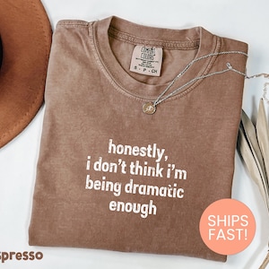 May include: Espresso-colored t-shirt with the text "honestly, I don't think I'm being dramatic enough" in white. A silver necklace with a pendant is draped across the shirt. The shirt is folded on a white surface.