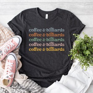 May include: A dark charcoal gray short-sleeve t-shirt featuring the repeated text "coffee & billiards" in five horizontal lines, each in a different color: light blue, orange, sage green, dusty pink, and mustard yellow, printed on the chest.