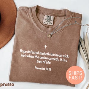 May include: A brown crew neck t-shirt featuring a white cross graphic and the text: Hope deferred maketh the heart sick: but when the desire cometh, it is a tree of life. Proverbs 13:12. The shirt has a Comfort Colors brand label at the inner neckline.
