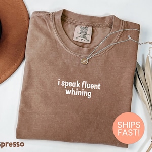 May include: Espresso-colored t-shirt with the phrase "i speak fluent whining" in white text. A silver necklace with a pendant rests on the shirt. The shirt is folded and displayed with a hat and dried plant.