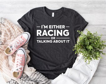 Puede incluir: Una camiseta gris oscuro de manga corta con el texto blanco "I'M EITHER RACING OR TALKING ABOUT IT" impreso en la parte delantera. La camiseta se muestra junto a un par de zapatillas de lona rosa claro, una bufanda beige y una pequeña planta verde.