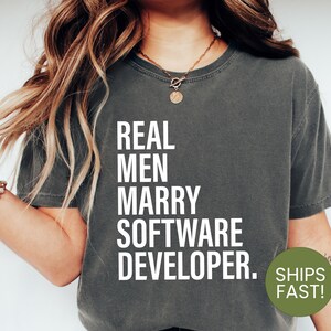 May include: A dark green t-shirt with white text that reads "REAL MEN MARRY SOFTWARE DEVELOPER." The text is centered on the shirt. The shirt is being worn by a person with long blonde hair.