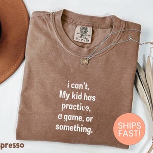 May include: Espresso-coloured t-shirt with the text "i can't. My kid has practice, a game, or something." A silver necklace with a pendant is draped on the shirt. The shirt is folded and has a Comfort Colors tag.