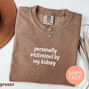 Peut inclure: T-shirt couleur expresso avec l'inscription blanche « personally victimized by my kidney ». Le t-shirt a un col rond et une petite étiquette. Un collier argenté avec un pendentif est sur le t-shirt.