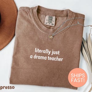 May include: Espresso-colored t-shirt with the text "literally just a drama teacher" in white. The shirt has a crew neck and short sleeves. A silver necklace with a pendant is draped on the shirt. A brown hat and dried leaves are in the background.
