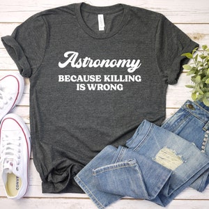 May include: A dark gray t-shirt with white text that reads "Astronomy because killing is wrong". The shirt is laid flat on a white wooden surface with a pair of white sneakers and a pair of blue jeans.