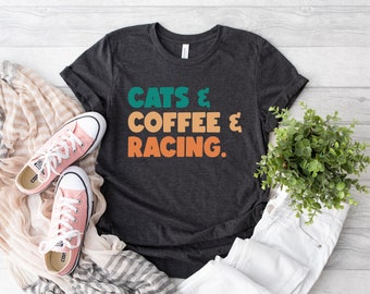 Puede incluir: Una camiseta de manga corta gris carbón oscuro con el texto "CATS & COFFEE & RACING." en letras apiladas, negritas y de estilo retro. La palabra "CATS" es de color verde azulado, mientras que "COFFEE" y "RACING." son de un tono naranja apagado.