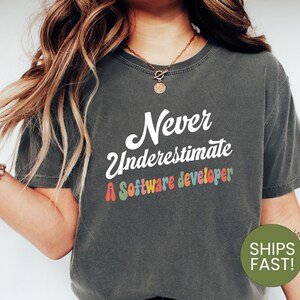 May include: A dark grey t-shirt with the text "Never underestimate a software developer" in white with colourful letters. The text is in a retro font style.