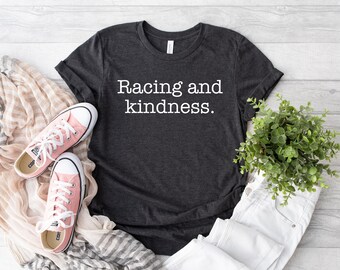 Puede incluir: Una camiseta gris oscuro de manga corta con el texto blanco "Racing and kindness." impreso en la parte delantera. La camiseta se muestra junto a un par de zapatillas de lona rosa claro, una bufanda a rayas beige y blanca y una pequeña planta verde.