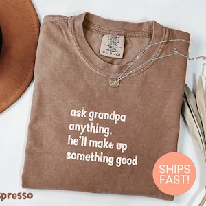 May include: Espresso-colored t-shirt with the text "ask grandpa anything. he'll make up something good" in white. A silver necklace with a pendant is draped on the shirt. The shirt is folded and displayed with a hat and dried plant.