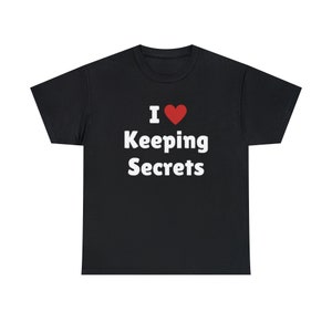 May include: A black short-sleeved t-shirt featuring the white text 'I' followed by a red heart symbol and the words 'Keeping Secrets' printed in a bold, sans-serif font centered on the chest area of the garment.