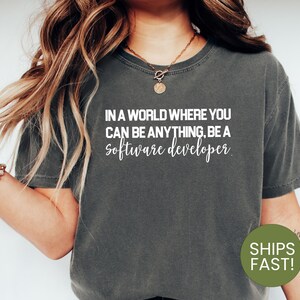 Peut inclure: Un t-shirt gris foncé avec du texte blanc qui dit "Dans un monde où vous pouvez être n'importe quoi, soyez un développeur logiciel."