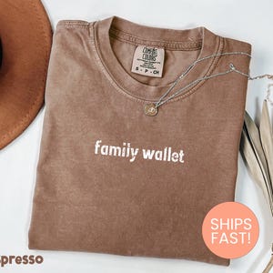 May include: Espresso-colored t-shirt with the words "family wallet" printed in white. A silver necklace with a pendant rests on the shirt. A brown hat and dried plant are in the background. The shirt has a Comfort Colors tag.